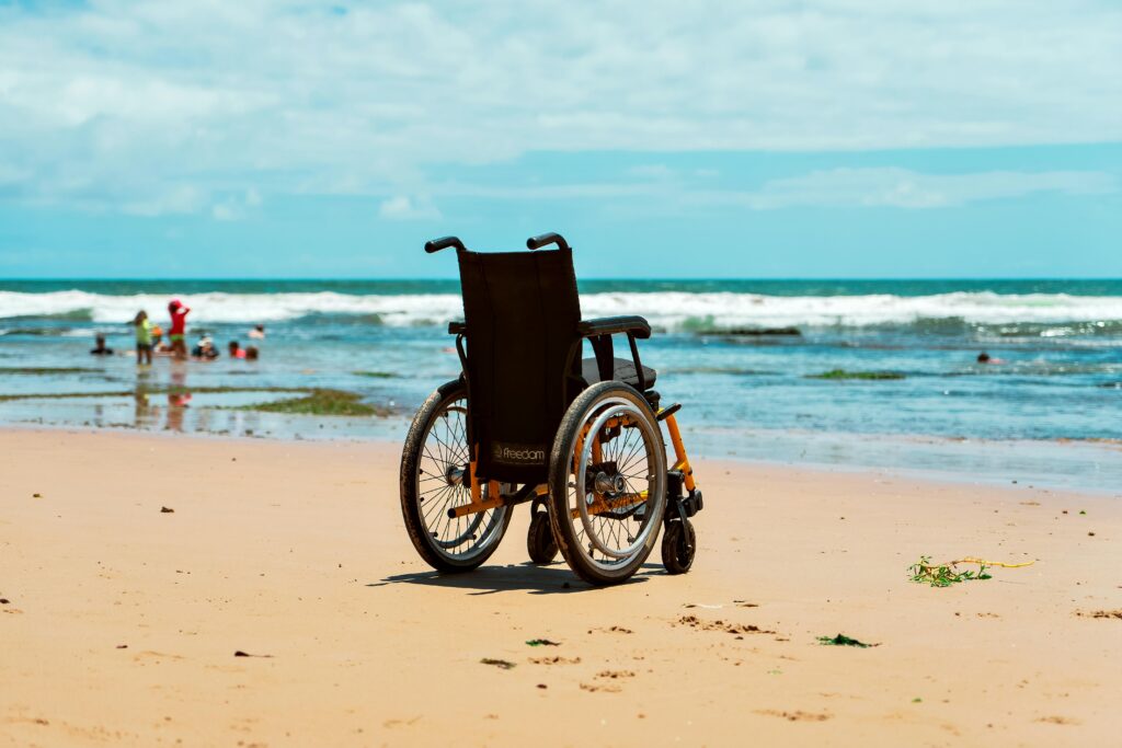 Wheelchair on sandy beach with ocean waves and distant people, highlighting accessibility and leisure.